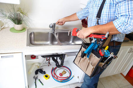 A handyman Mandurah worker fixing a leaking sink tap with a tool belt on and his tool laid out around the sink floor