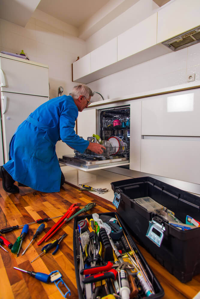 A senior handyman Mandurah worker fixing a broken dishwasher in blue overalls with his toolbox open next to the dishwasher full of tools