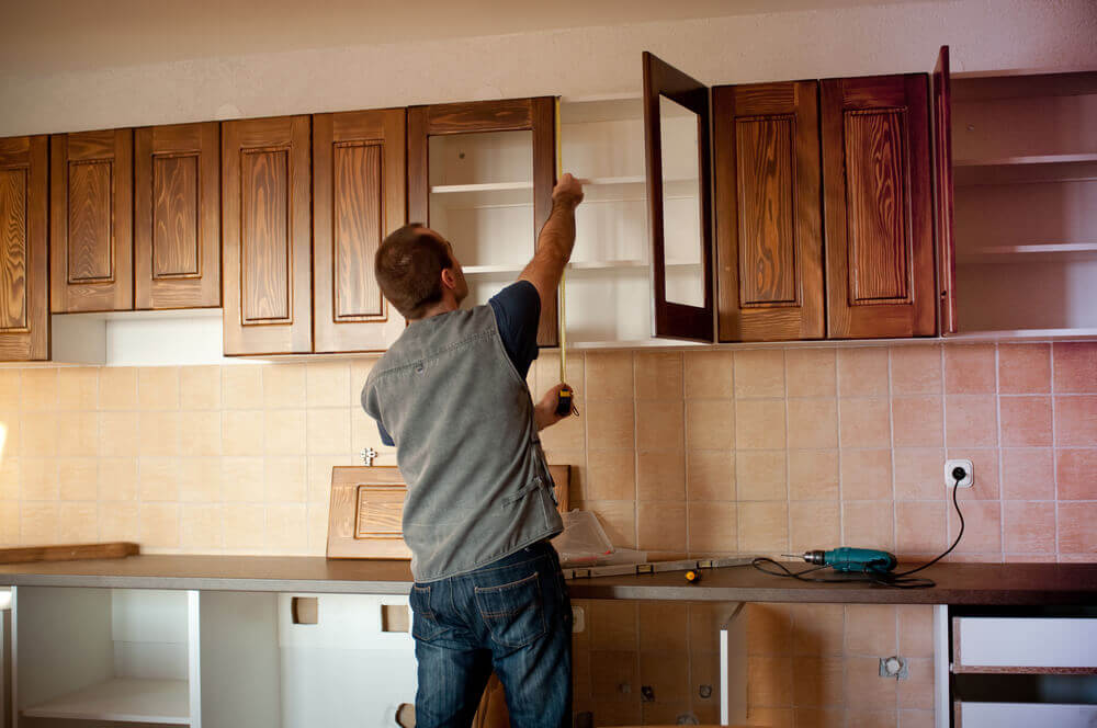A handyman Mandurah worker installing some new dark brown kitchen cabinets whilst checking a measurement with a tape measure  