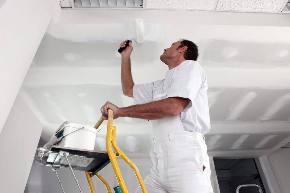 A handyman Mandurah worker in some white overalls painting a ceiling on a ladder with a paint roller