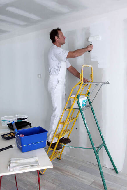 A painters Mandurah worker standing on a ladder painting the top of a white wall in whit overalls and his paint on the table 
