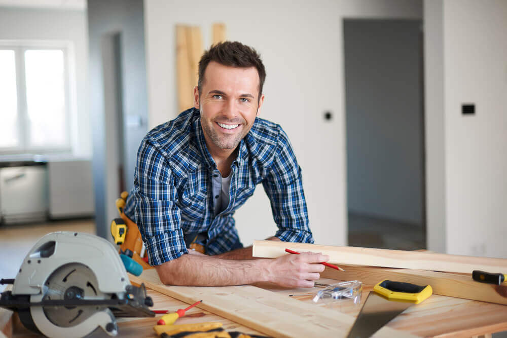 A carpentry Mandurah worker leaning on a table in a house smiling with his tools and some half made wooden structures near him